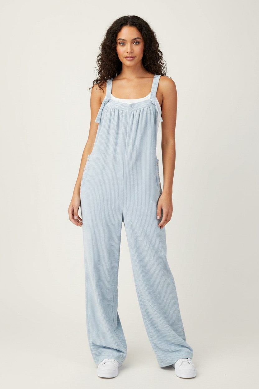 A woman modeling a light blue ribbed wide-leg jumpsuit with white trim on the neckline and shoulder straps, paired with white sneakers, against a plain light gray studio background.