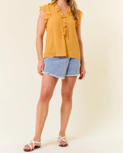 Woman wearing a yellow ruffled top and denim shorts against a plain background.