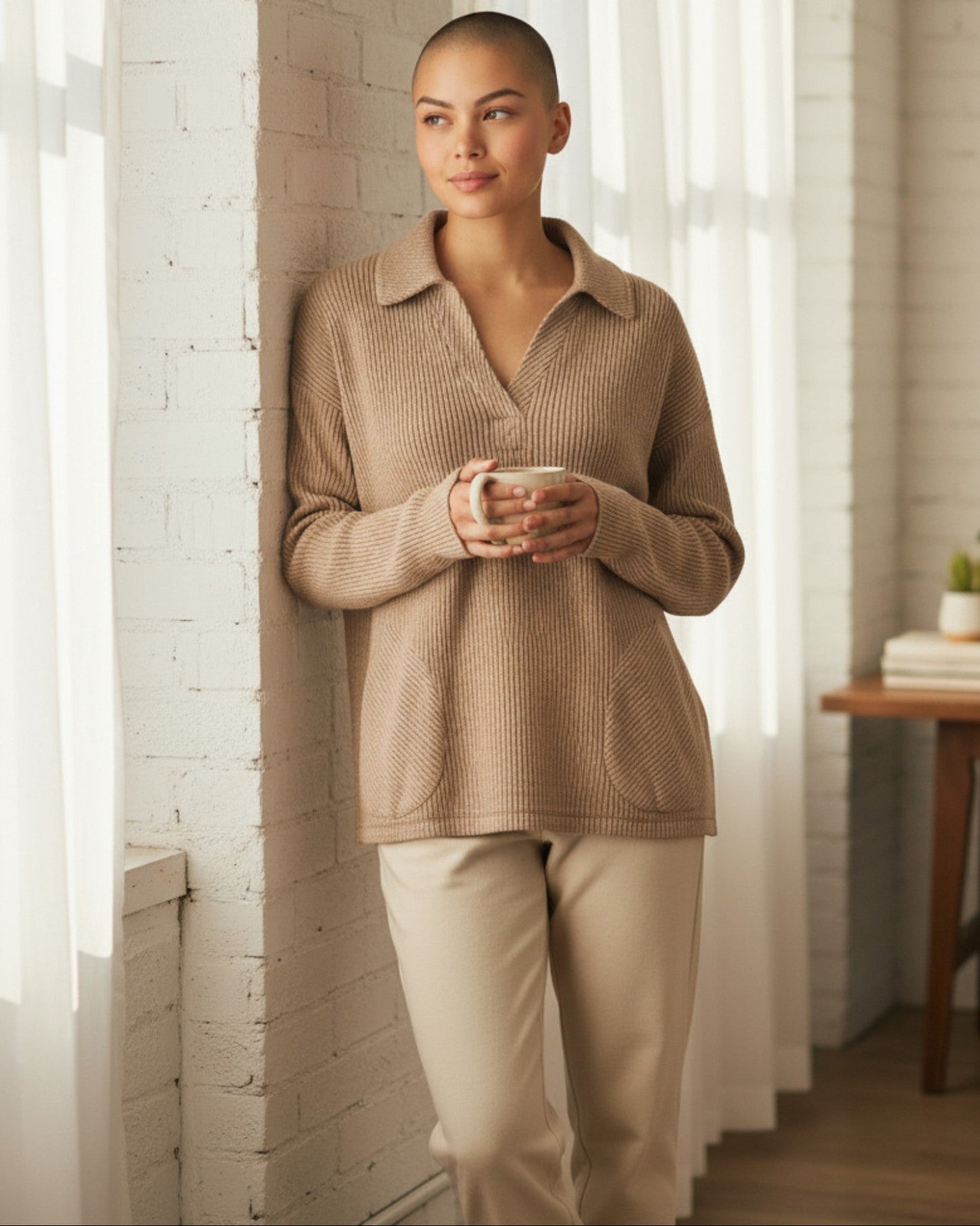 Woman in a acorn colored ribbed sweater and pants holding a mug, standing in a bright room with white walls and a wooden table.