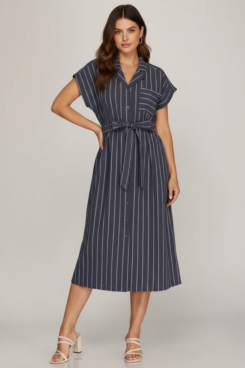 A woman modeling a navy blue midi shirt dress with thin white vertical pinstripes, featuring short sleeves, a button-down front, and a matching tie belt at the waist, set against a neutral light gray background.