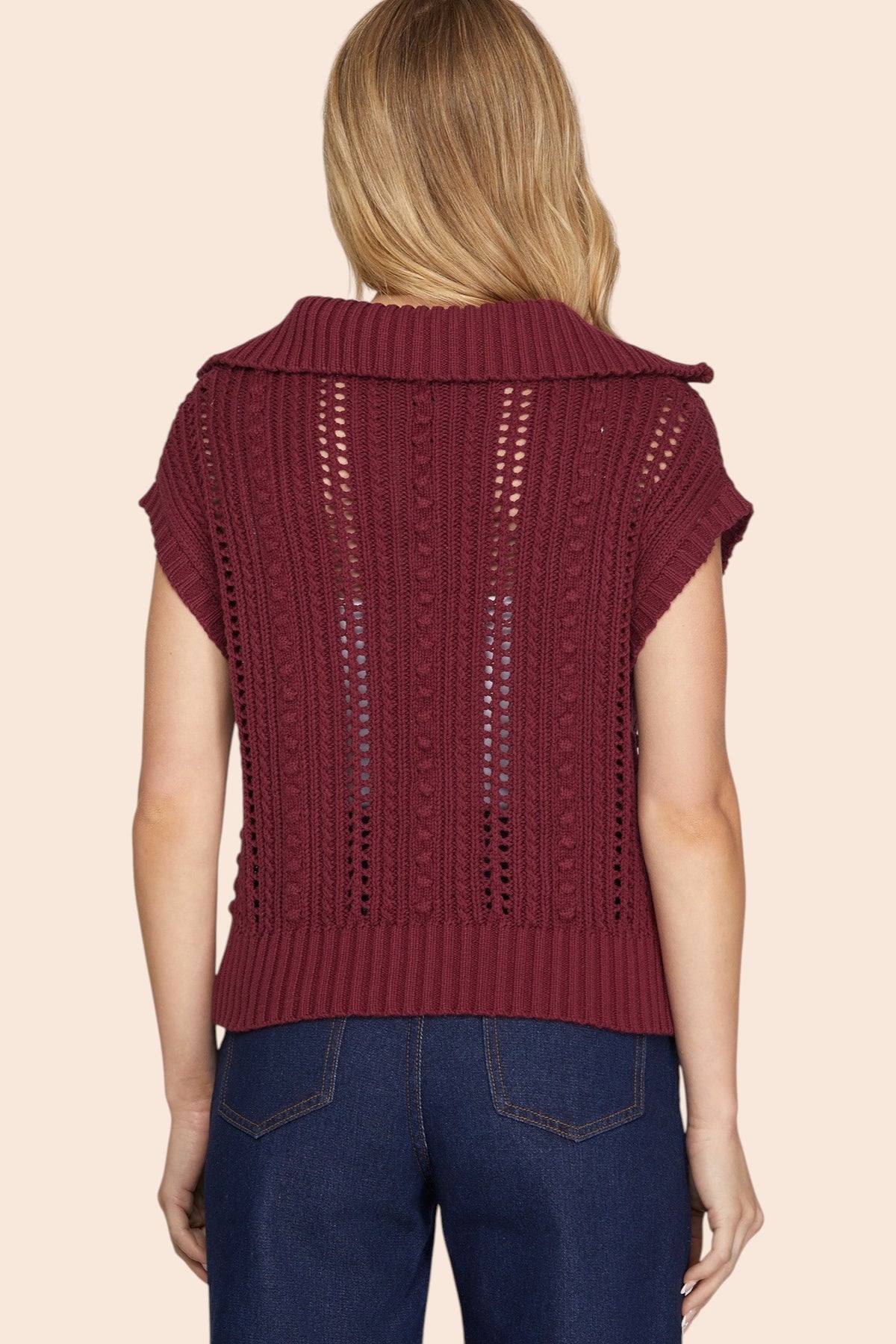 A woman viewed from the back wearing a burgundy open-knit cropped cap sleeve sweater  with short sleeves and a wide collar, paired with dark blue jeans, against a solid light beige background.