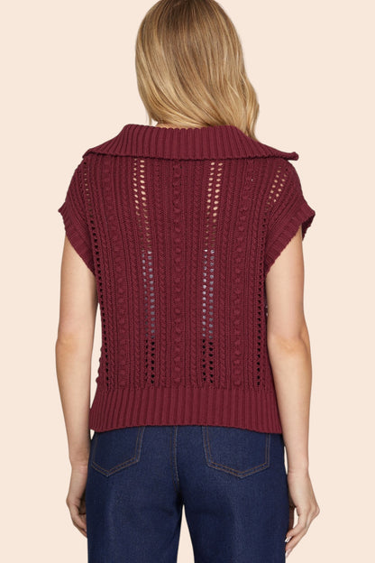 A woman viewed from the back wearing a burgundy open-knit cropped cap sleeve sweater  with short sleeves and a wide collar, paired with dark blue jeans, against a solid light beige background.