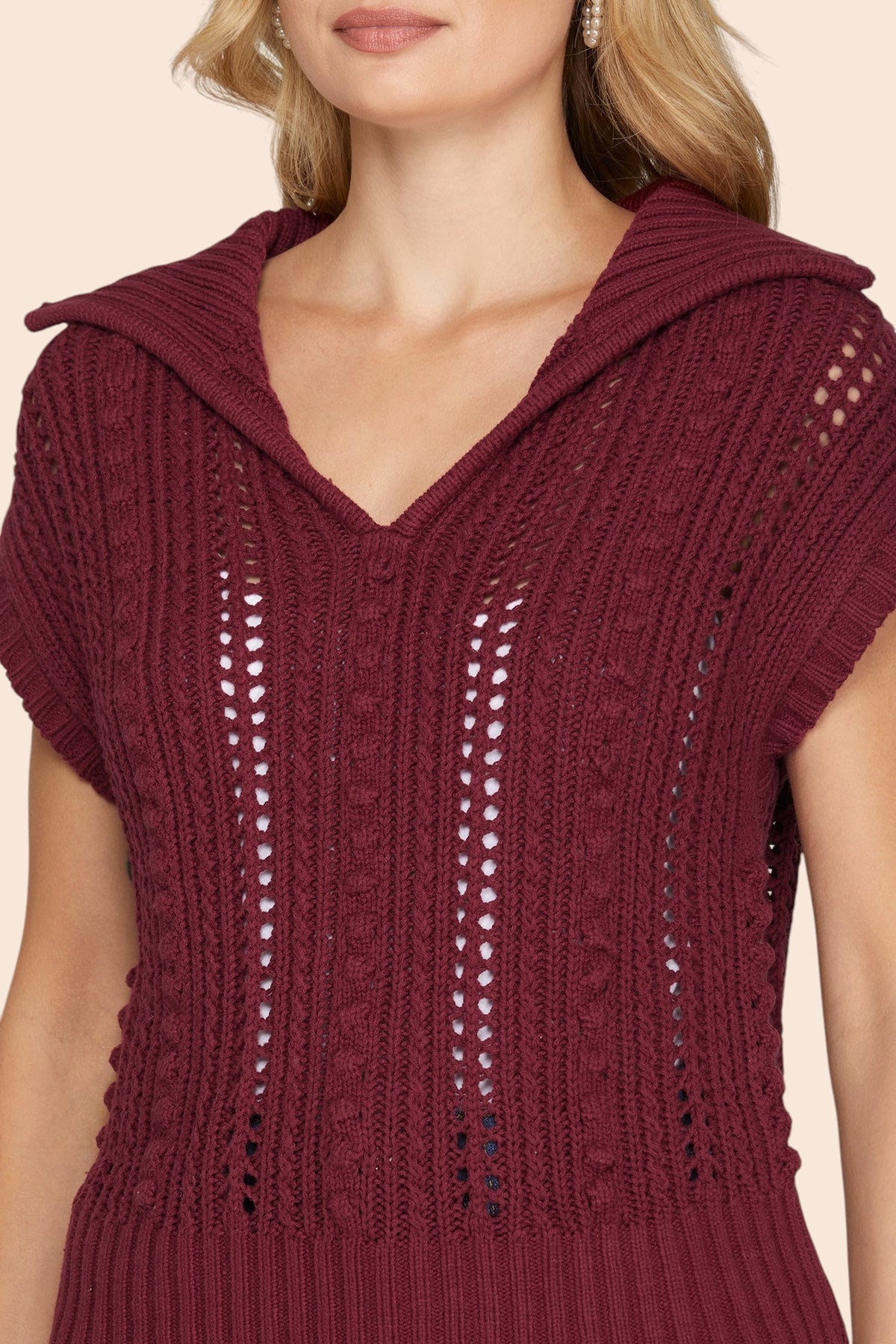 A close-up of a woman wearing a burgundy knit sweater with short sleeves and openwork vertical details, shown against a light beige background. 