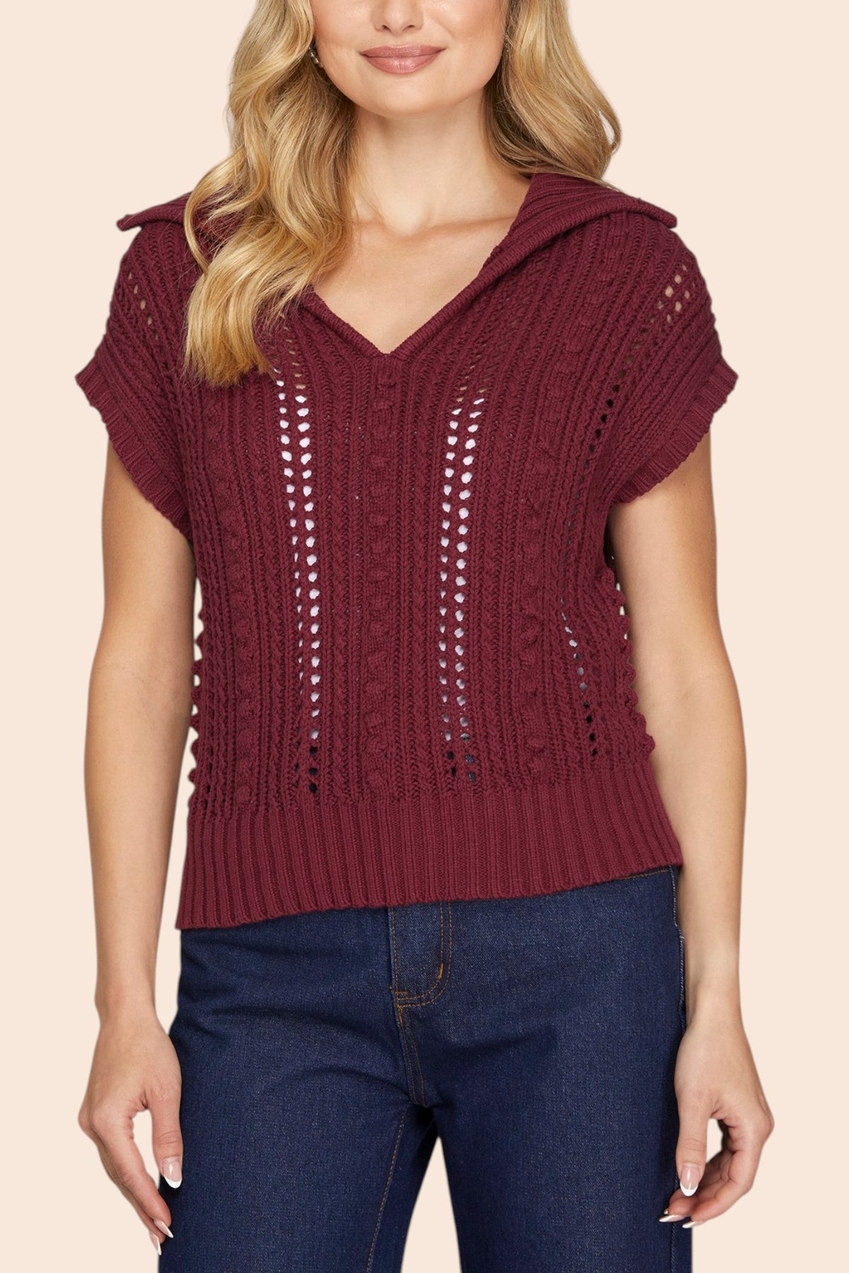 A woman wearing a burgundy knit short-sleeve top with a V-neck and textured openwork details, paired with dark blue jeans, against a solid light beige background.