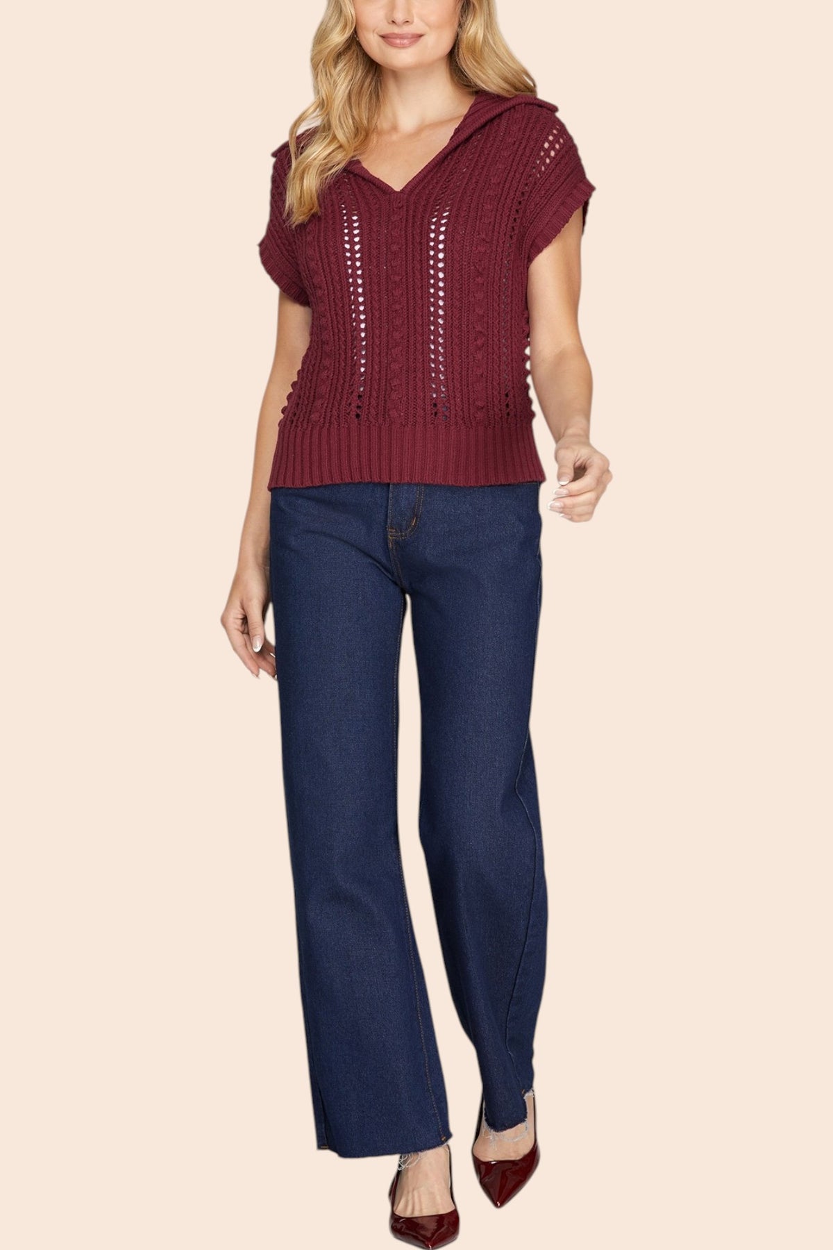 A woman modeling a burgundy short-sleeve knit top with a V-neck and textured knit pattern, paired with dark blue jeans and burgundy pointed-toe heels, against a solid beige background.