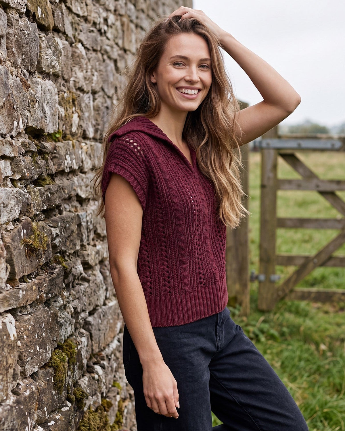 A woman stands outdoors against a stone wall wearing a sleeveless ribbed knit top in deep burgundy and dark navy pants.