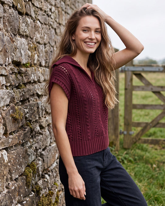 A woman stands outdoors against a stone wall wearing a sleeveless ribbed knit top in deep burgundy and dark navy pants.