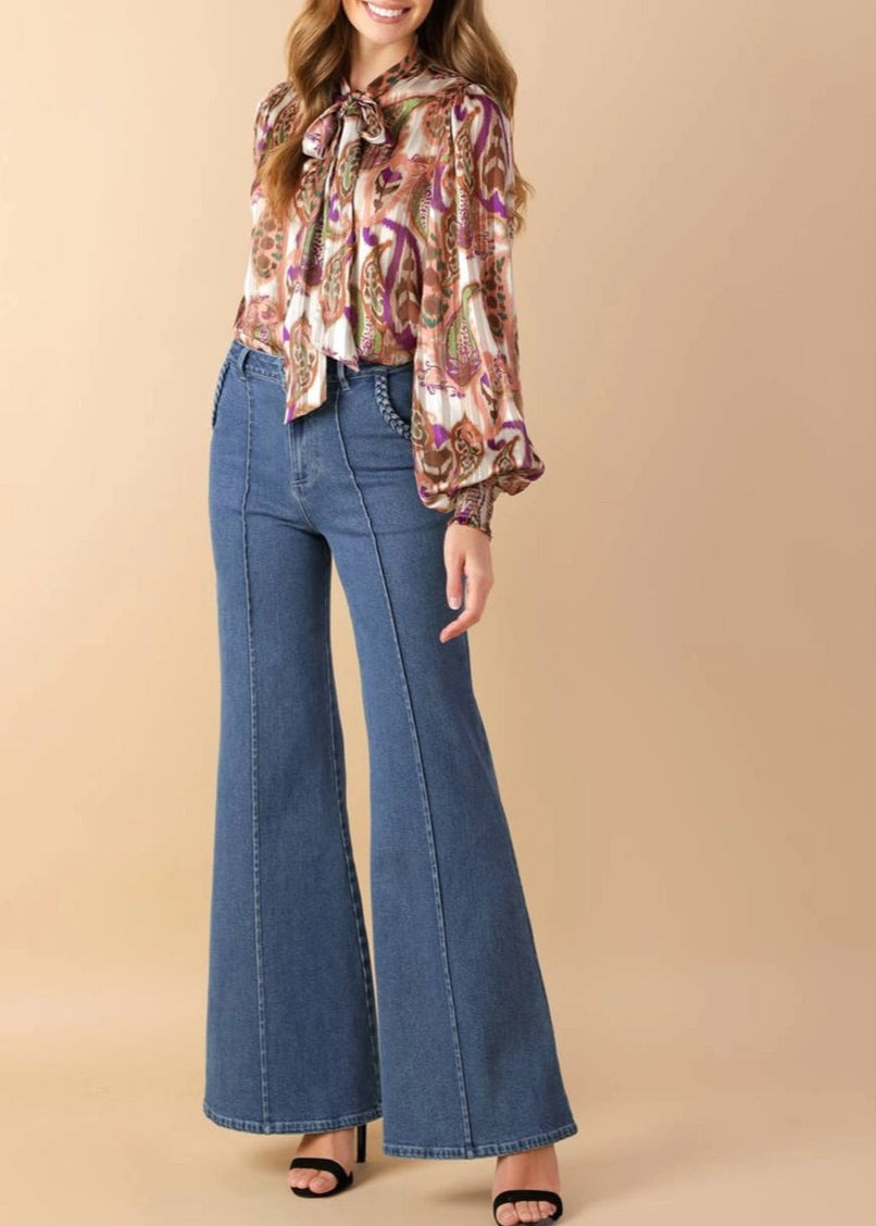 Woman wearing a patterned blouse and blue flared jeans on a beige background.