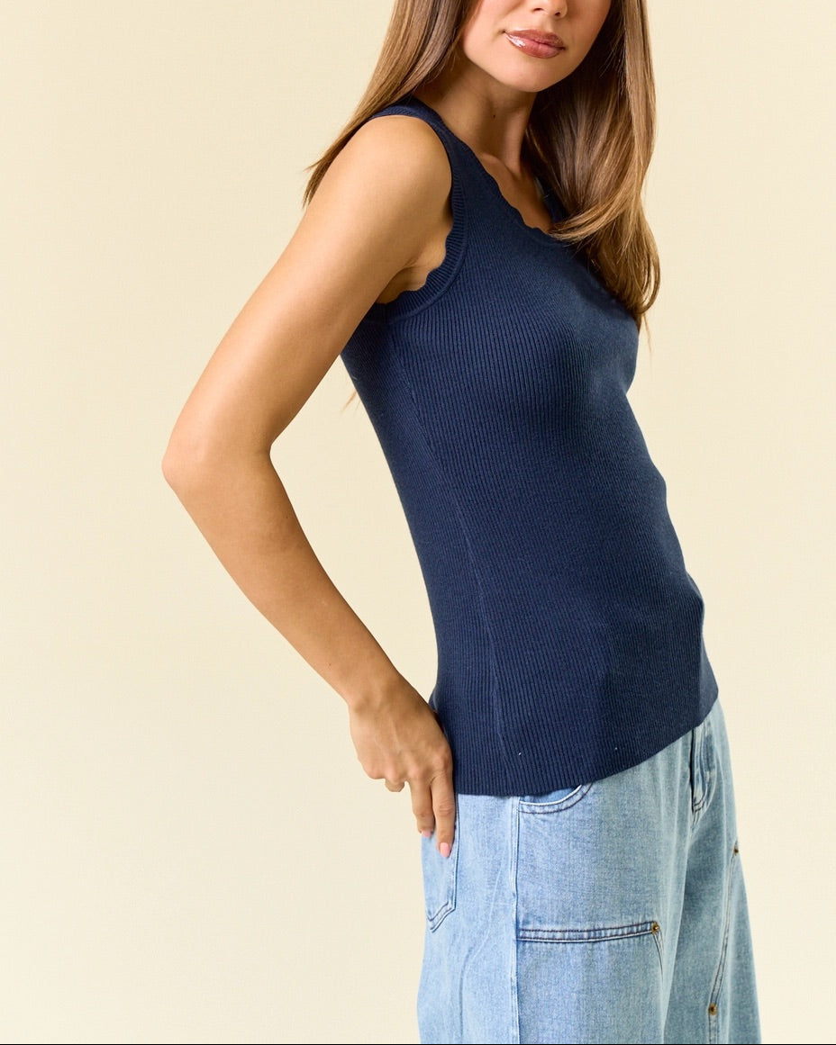 Woman wearing a navy blue sleeveless scalloped top and light blue jeans against a beige background.