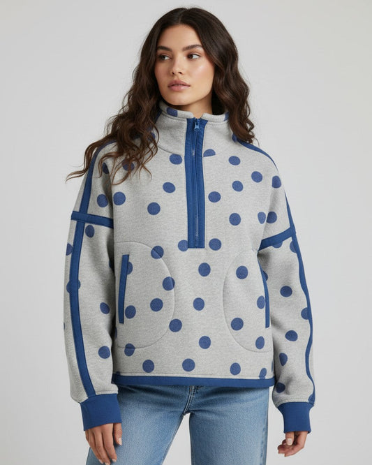 A woman modeling a light gray heathered quarter-zip sweatshirt with blue polka dots and solid blue trim, paired with light blue jeans, against a light gray studio background.