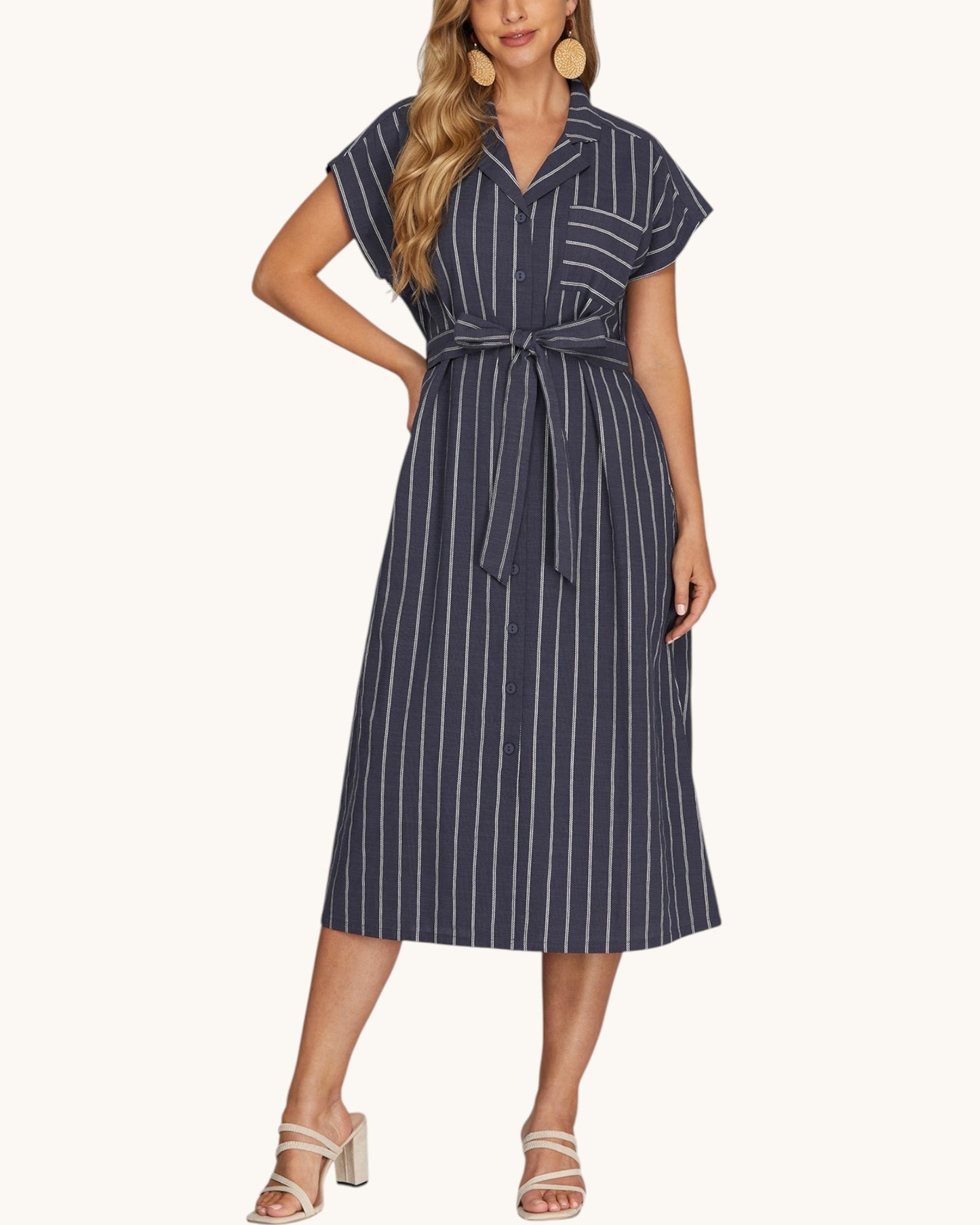 A woman modeling a navy blue midi shirt dress with white vertical pinstripes, featuring short sleeves, a collared neckline, and a matching waist tie, paired with beige heeled sandals against a light cream background.
