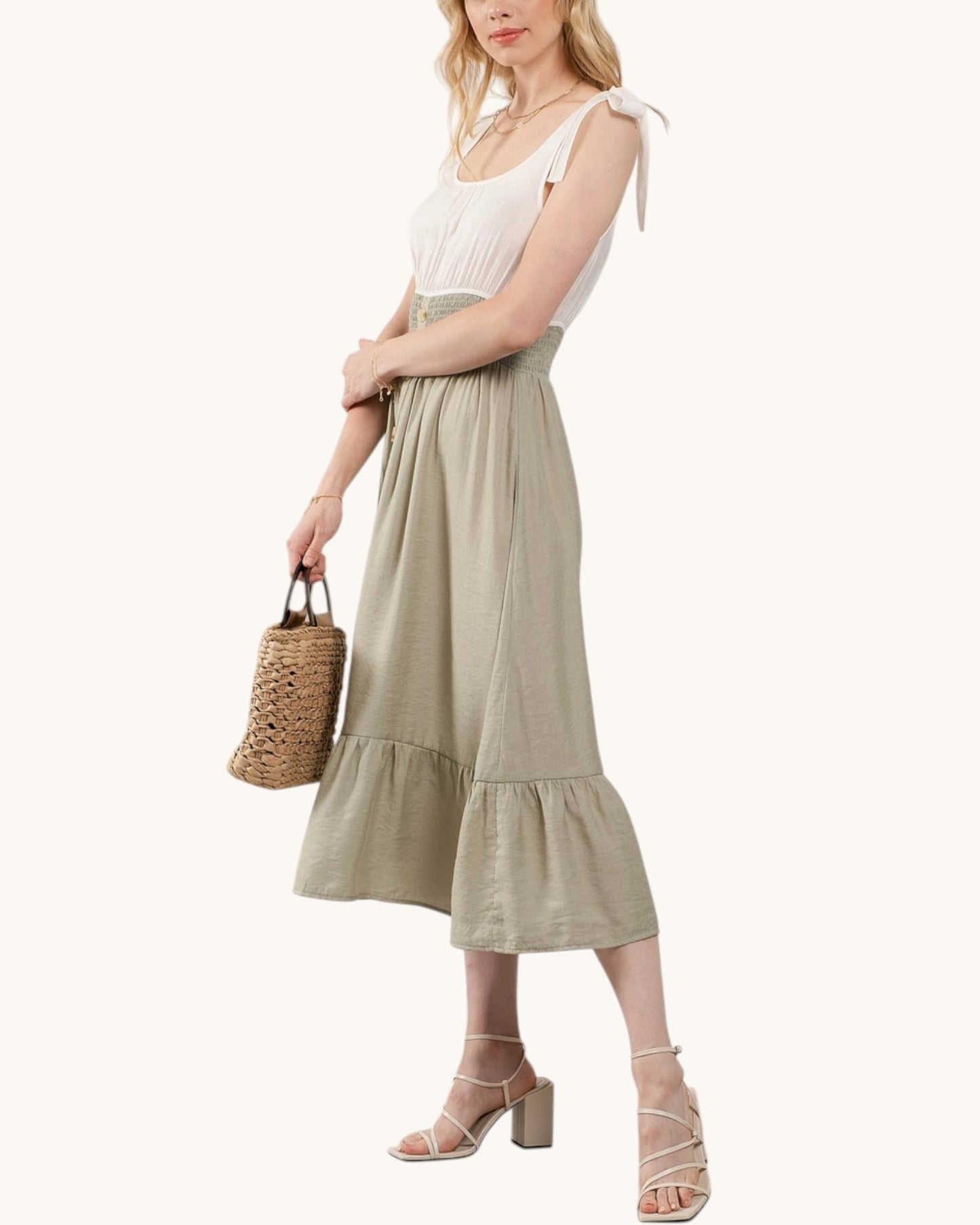 Woman wearing a dress with a contrasting white top and sage skirt holding a woven bag on a white background.