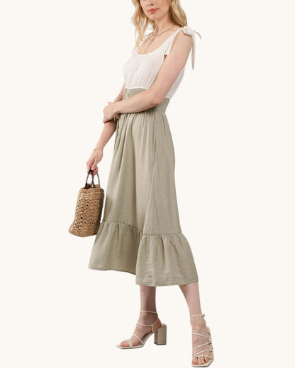 Woman wearing a dress with a contrasting white top and sage skirt holding a woven bag on a white background.