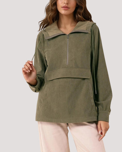 A woman wearing an olive green corduroy velour half-zip pullover jacket with a wide collar and drawstring details, paired with light pink pants, against a solid light gray background.