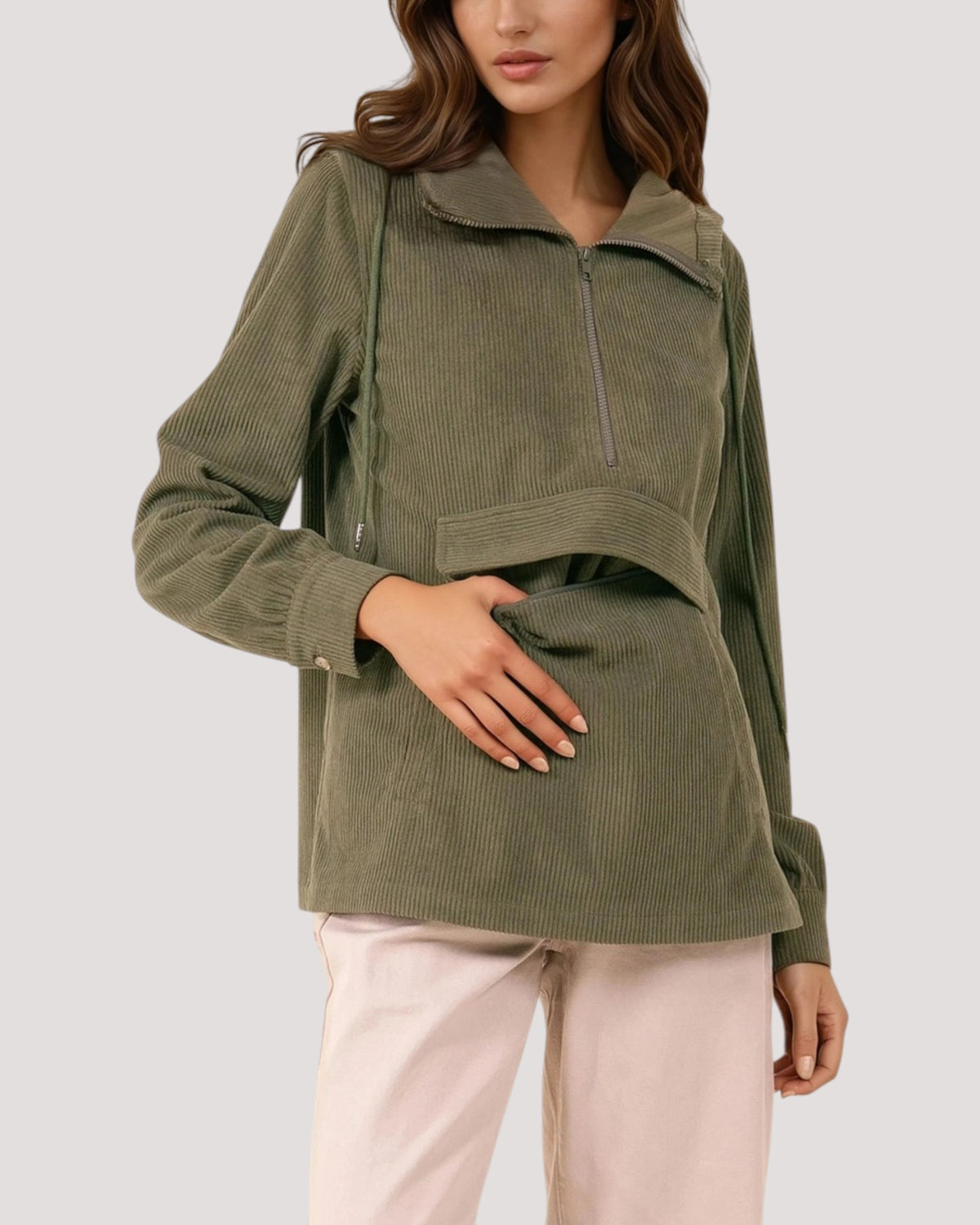 A woman wearing an olive green corduroy half-zip pullover with a high collar and drawstring details, paired with light pink pants, against a solid light gray background.