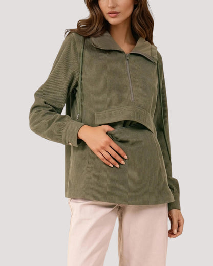 A woman wearing an olive green corduroy half-zip pullover with a high collar and drawstring details, paired with light pink pants, against a solid light gray background.