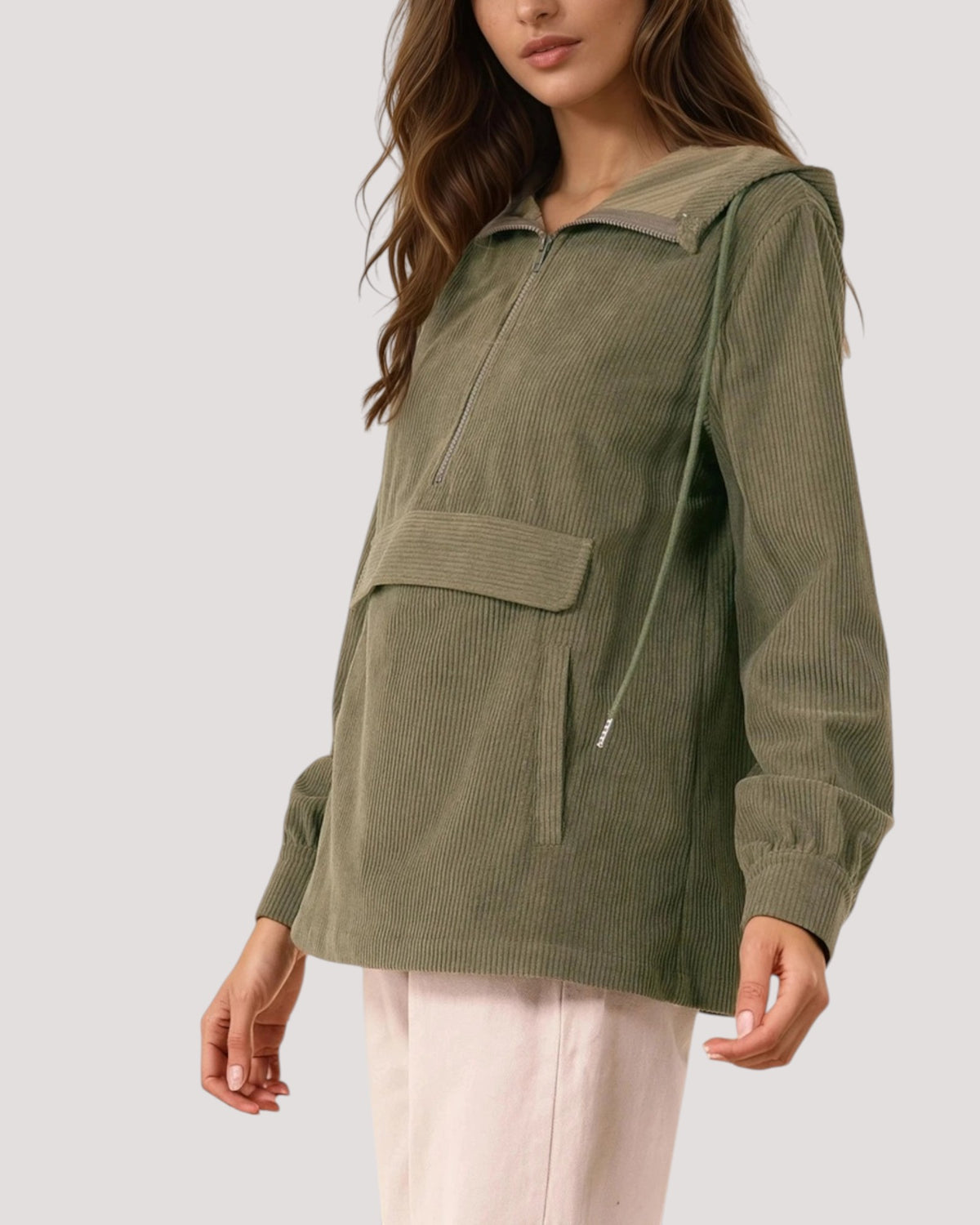 A woman wearing an olive green corduroy hooded pullover jacket with a half-zip front and drawstring, paired with light pink pants, against a solid light gray background.