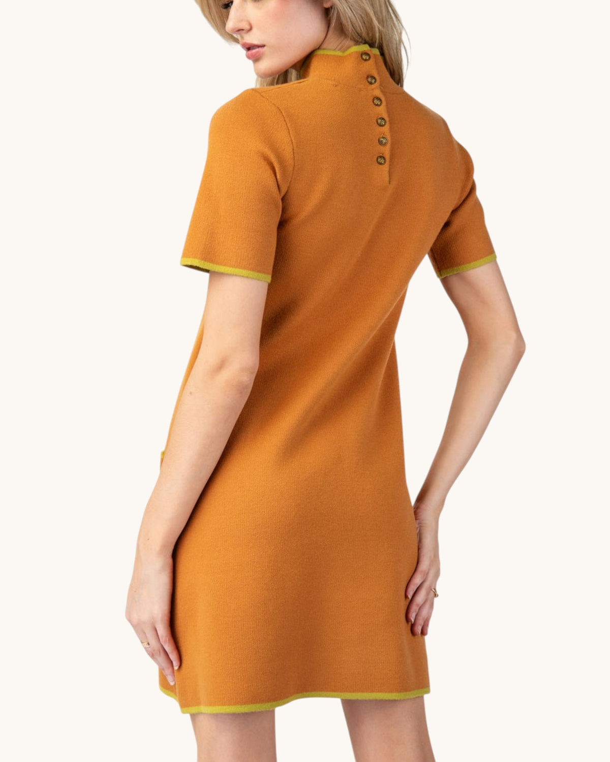A woman modeling a short-sleeve orange knit dress from the back, featuring a high mock neck with decorative buttons and contrasting light green trim on the sleeve cuffs and hem.