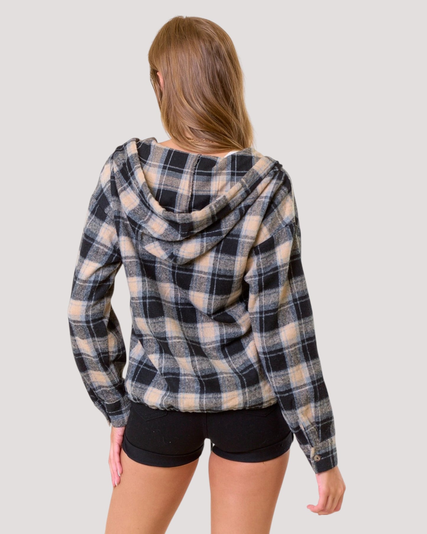 A woman shown from the back wearing a black, beige, and light blue plaid hooded sweatshirt and black shorts against a solid light gray background.