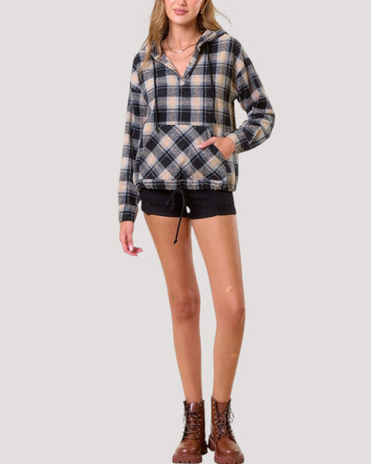 A woman modeling a long-sleeve plaid hooded pullover top with black shorts and brown lace-up boots, photographed against a plain light gray studio background.