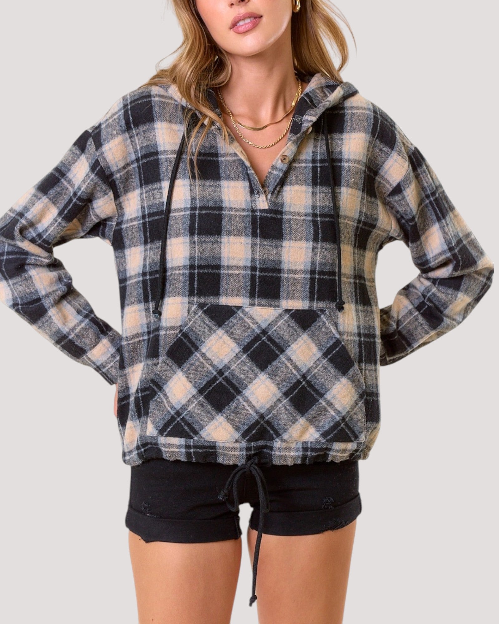A woman wearing a black, beige, and light blur plaid pullover hoodie with a half-zip front and drawstring hood, paired with black shorts, against a solid light gray background.