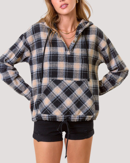 A woman wearing a black, beige, and light blur plaid pullover hoodie with a half-zip front and drawstring hood, paired with black shorts, against a solid light gray background.