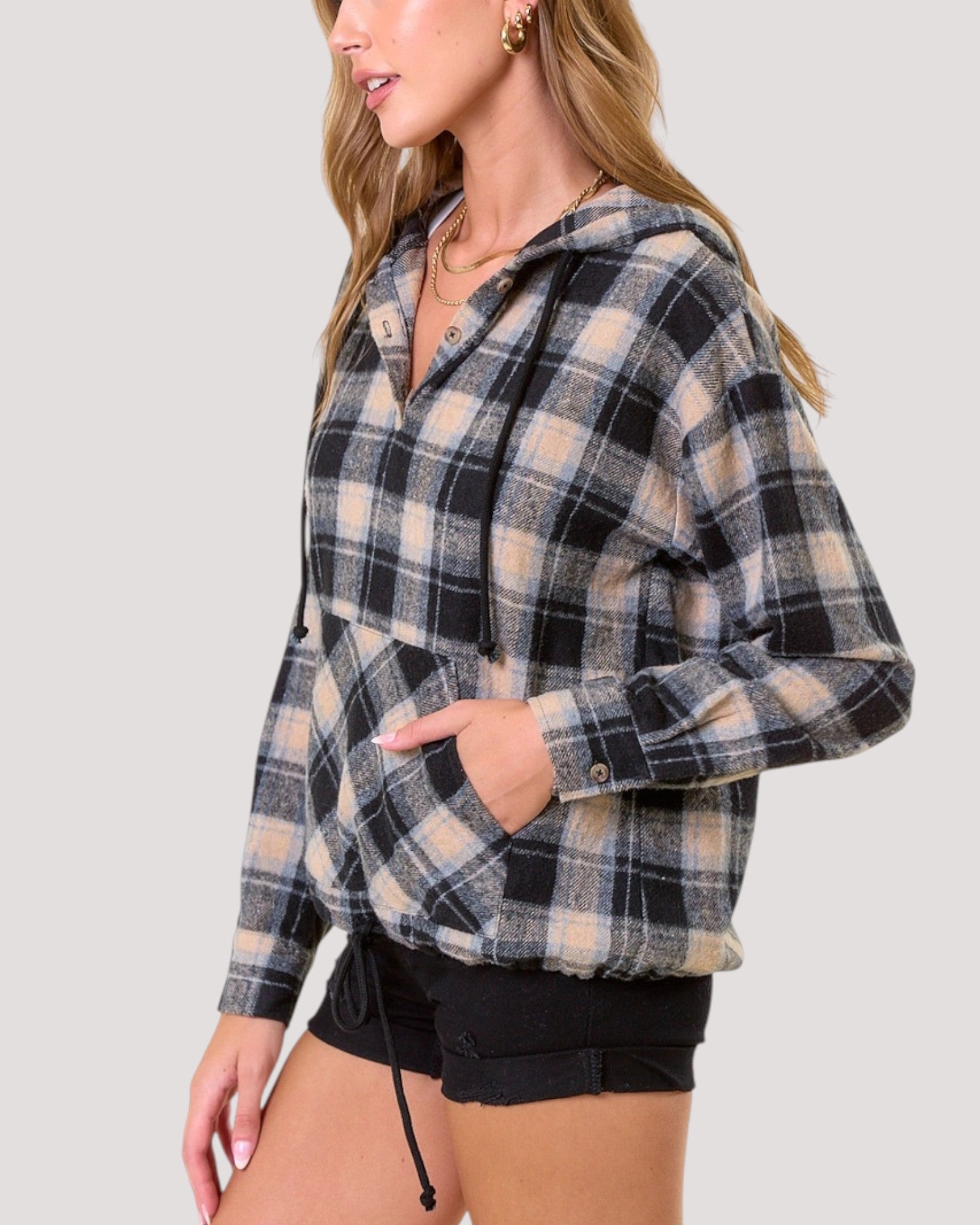 A woman wearing a black, beige, and light blue plaid hooded pullover sweatshirt with a half-button placket and front pockets, paired with black shorts, against a solid light gray background.