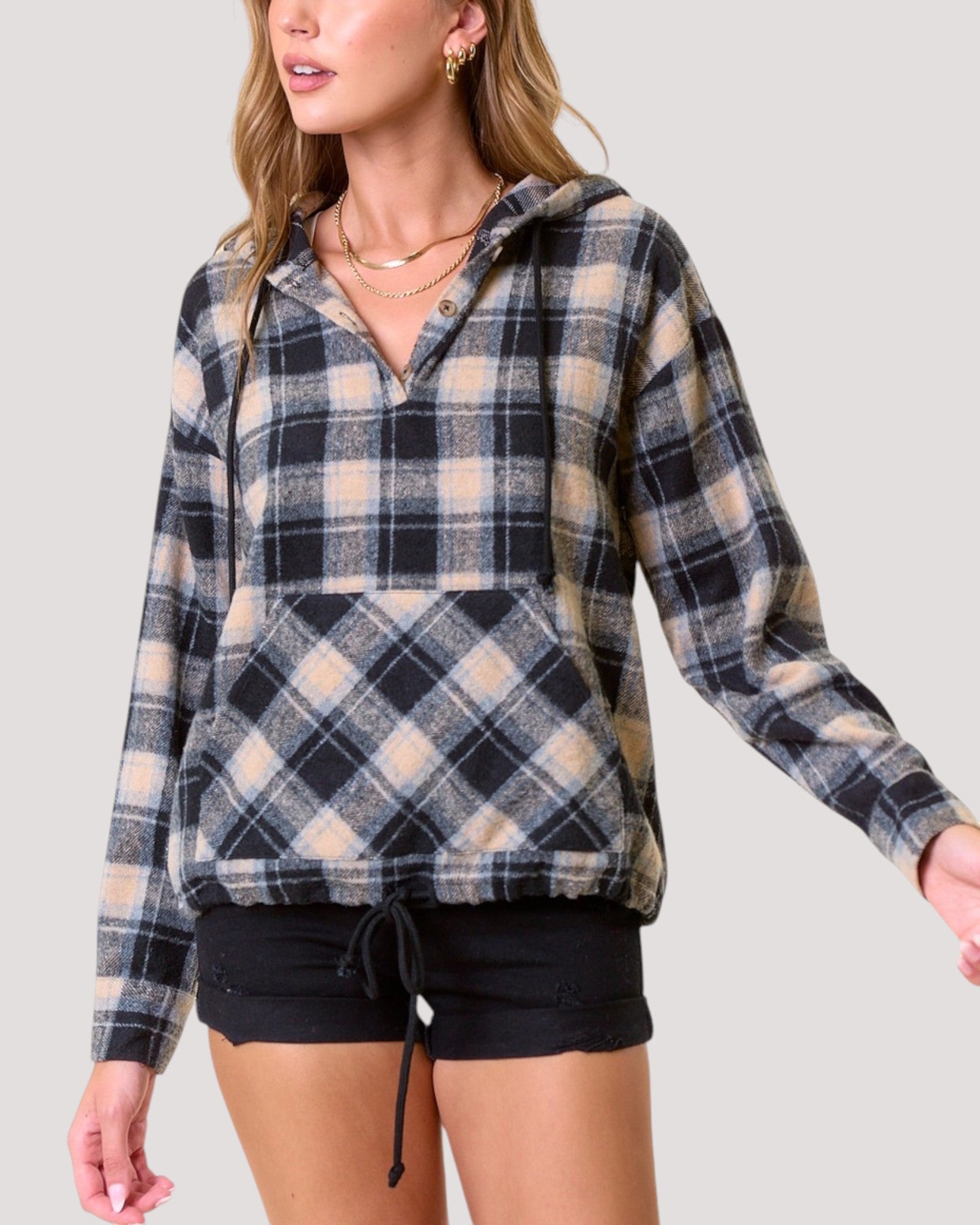 A woman wearing a long-sleeve hooded pullover sweatshirt in a beige, black, and light blue plaid pattern, paired with black shorts, against a solid light gray background.