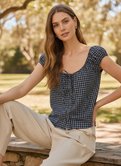 A woman sitting outdoors on a bench wearing a navy plaid short-sleeve top with a scoop neckline and tie detail, paired with beige pants.