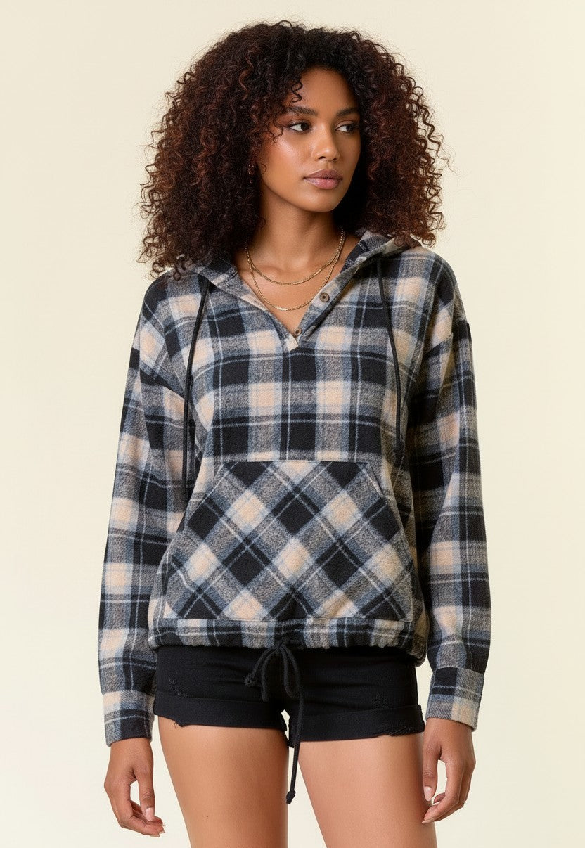 A woman with curly brown hair wearing a black, beige, and light blue plaid pullover hoodie with a half-button placket and a front kangaroo pocket, paired with black shorts, against a solid cream background.