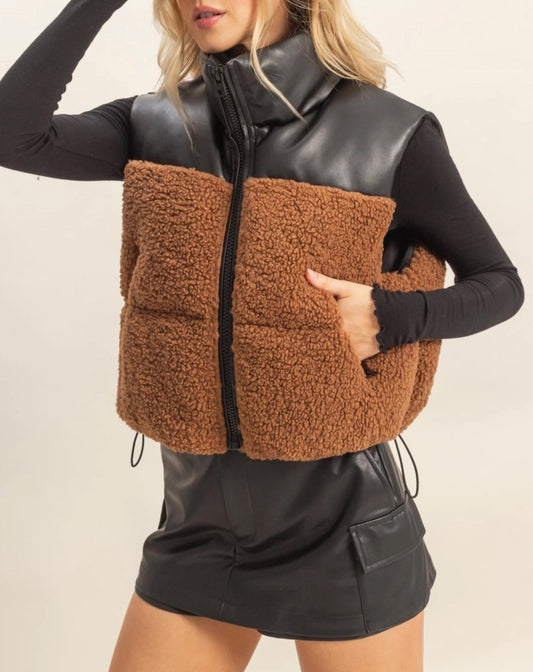 Woman wearing a brown sherpa and black faux leather vest with a plain background.