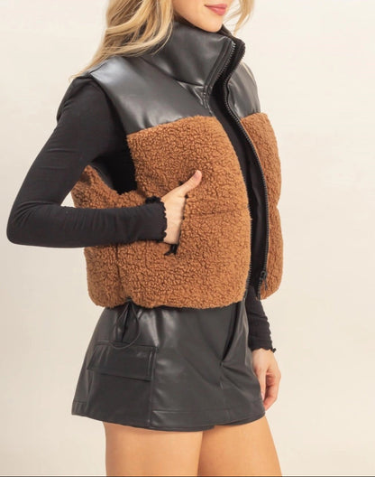 Woman wearing a brown sherpa and black faux leather vest with a plain background.