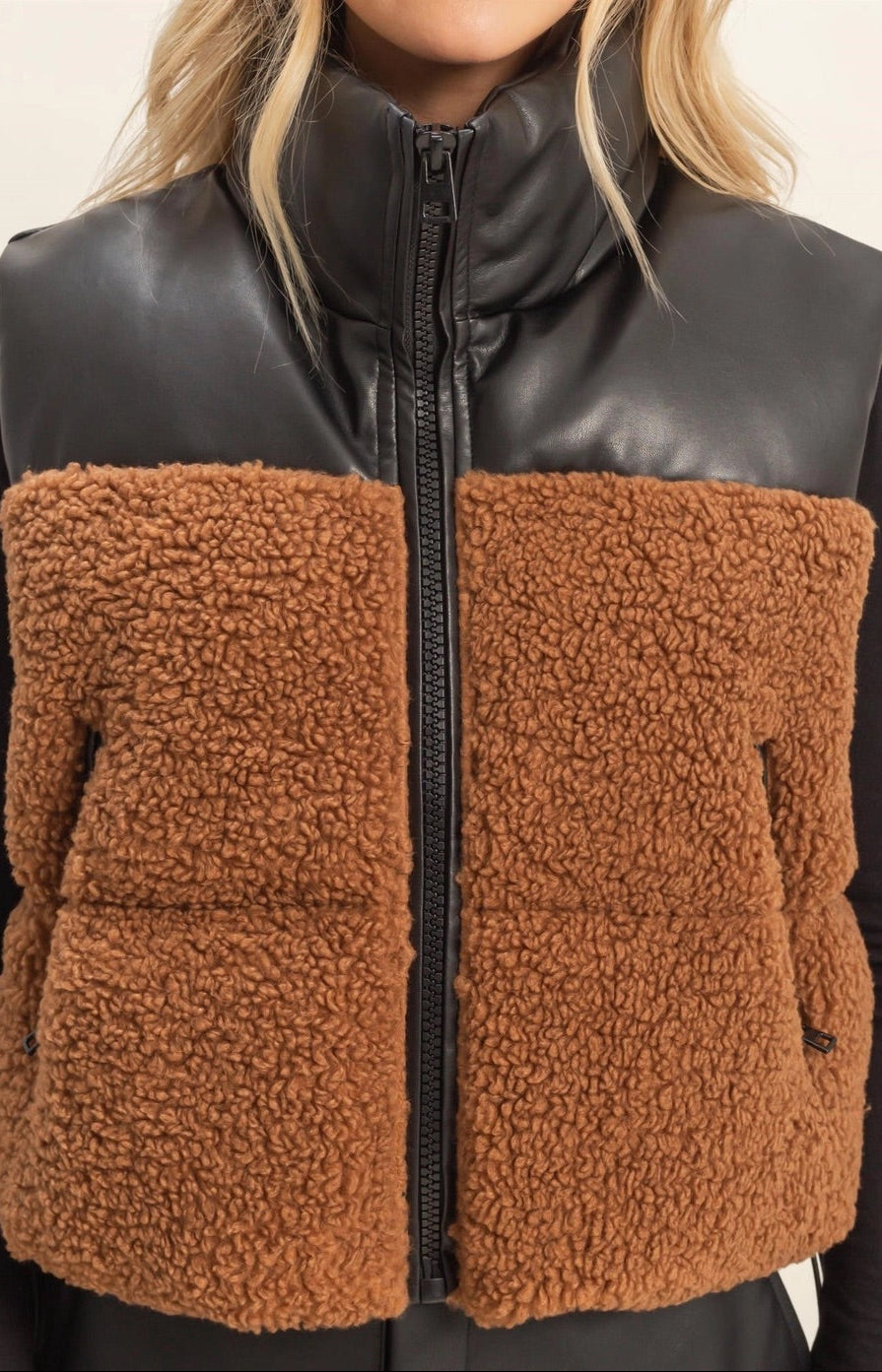 Woman wearing a brown sherpa and black faux leather vest with a plain background.
