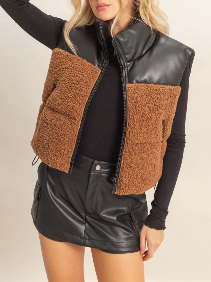 Woman wearing a brown sherpa and black faux leather vest with a plain background.