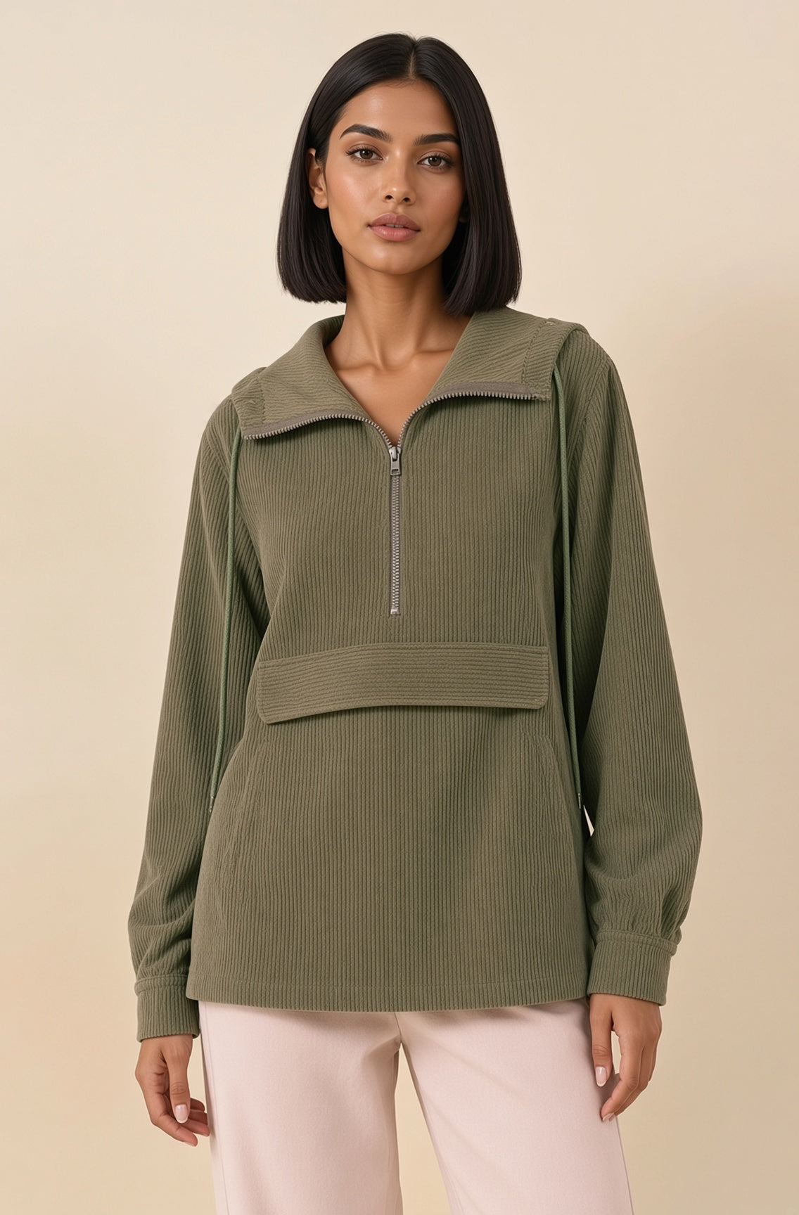 A woman modeling a ribbed olive green half-zip pullover with a mock neck and front patch pocket, paired with light pink pants, against a solid beige background.