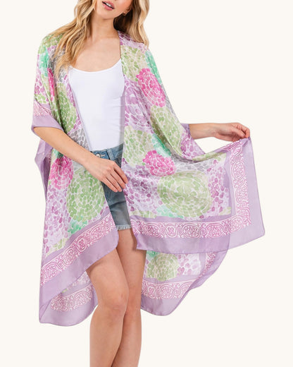 Woman wearing a green, pink and purple floral kimono over a white top and denim shorts on a white background.