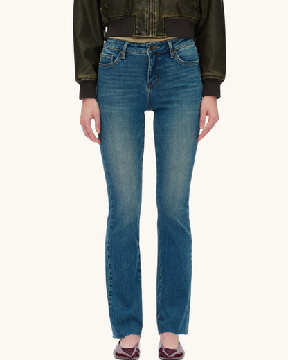 A model wearing a pair of medium-wash blue denim jeans with a straight leg with raw hem, styled with a leather jacket and burgundy pointed flats against a plain off-white background.