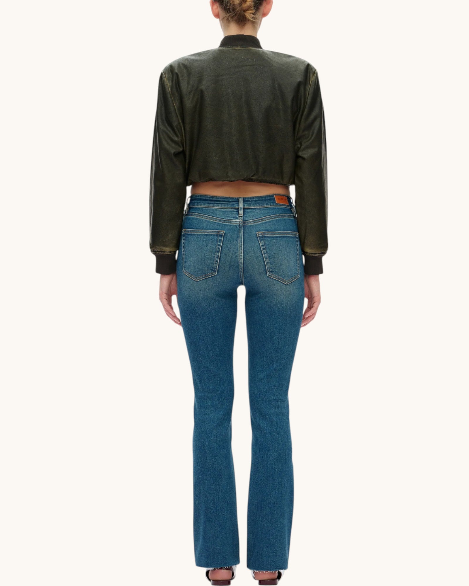 A model shown from the back wearing a dark olive green cropped leather jacket and vintage blue wash straight jeans with raw hem, set against a plain off-white background.