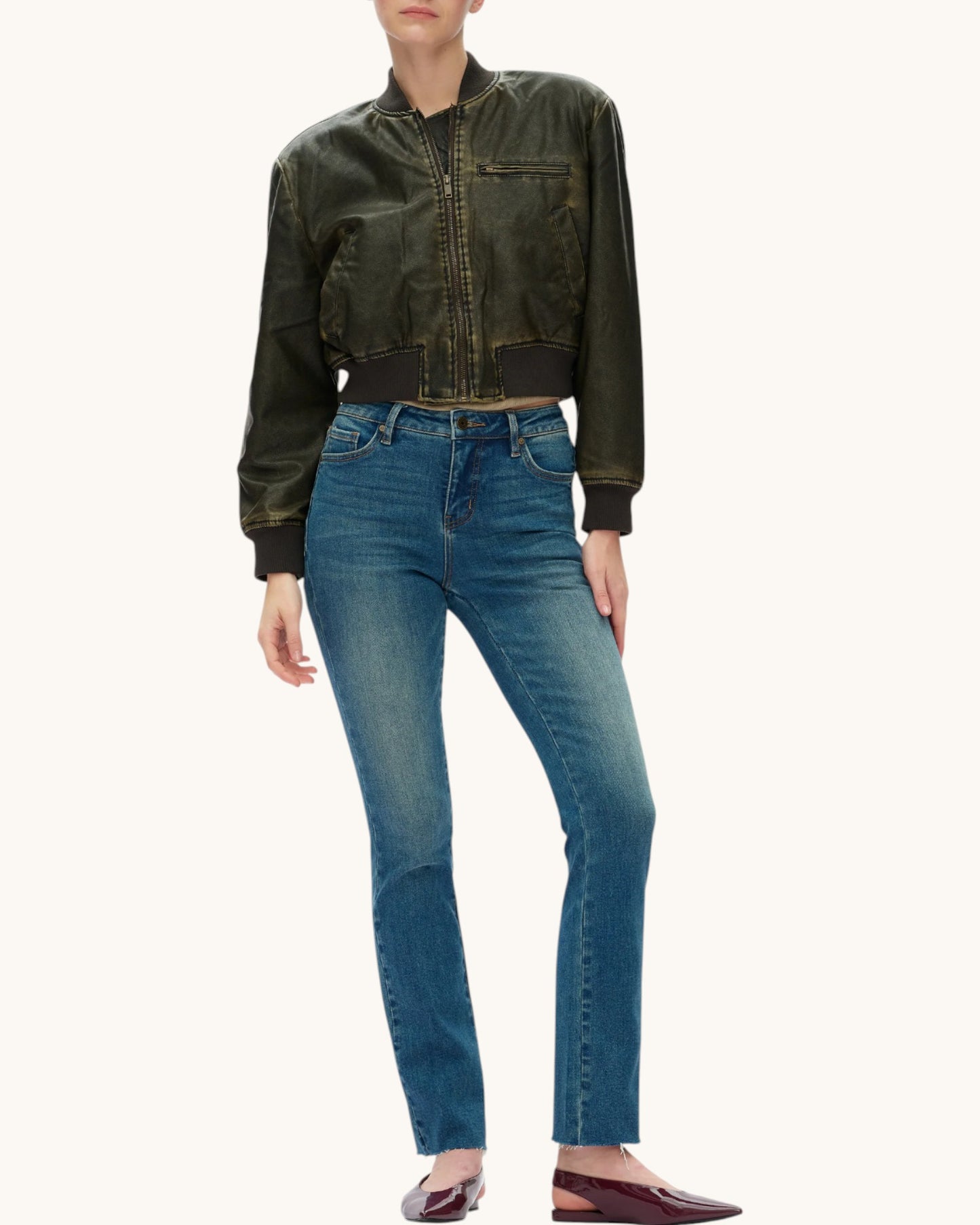 A model wearing a dark olive green cropped leather bomber jacket and blue jeans, paired with burgundy pointed flats, against a solid off-white background.