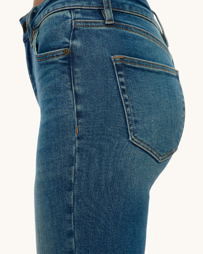 Close-up of the back waist and upper legs of a person wearing medium blue denim jeans, showing the back pocket, stitching, and waistband against a plain light background.