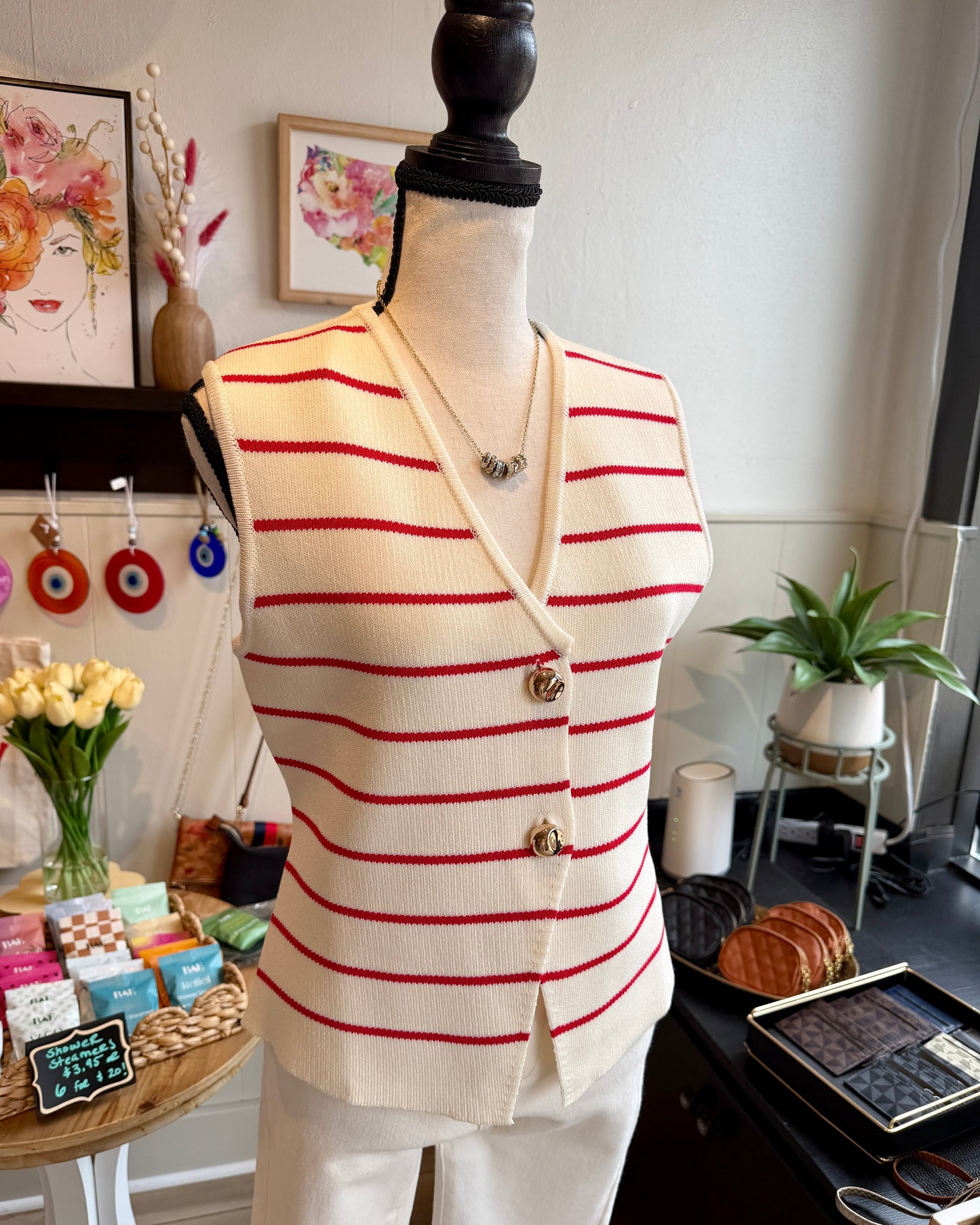 A cream knit vest with thin red horizontal stripes and gold buttons, displayed on a mannequin torso in a boutique setting.