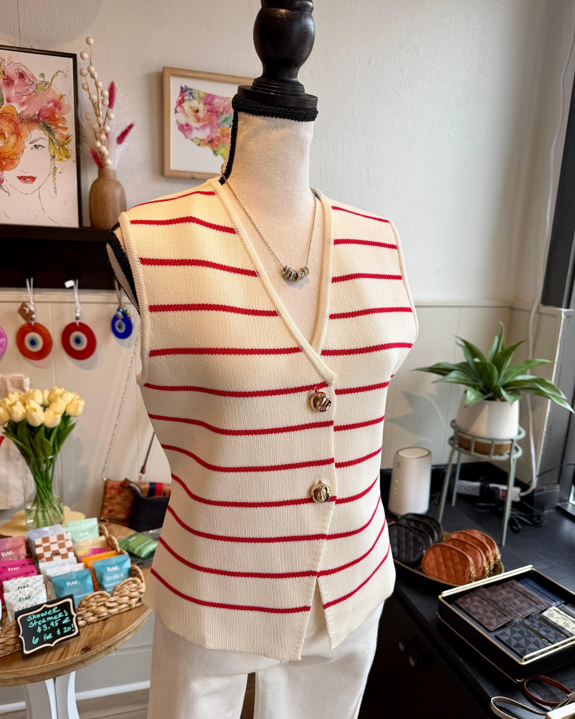 A cream knit vest with thin red horizontal stripes and gold buttons, displayed on a mannequin torso in a boutique setting.