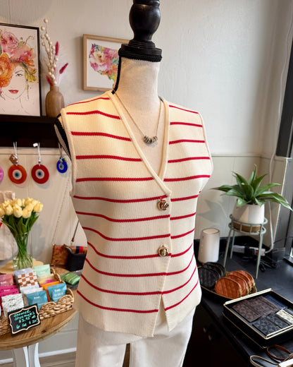 A cream knit vest with thin red horizontal stripes and gold buttons, displayed on a mannequin torso in a boutique setting.