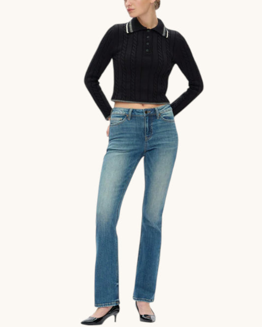 A model wearing a black long-sleeve knit polo crop top with white trim on the collar and button placket, paired with medium wash bootcut blue jeans and black pointed-toe heels, against a solid off-white background.