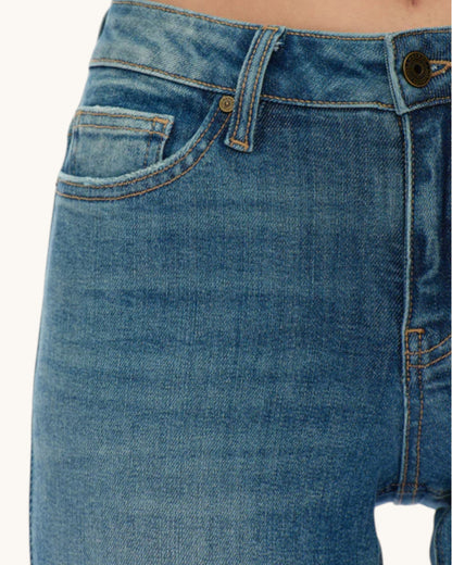Close-up of blue denim jeans showing the front pocket, waistband, button, and stitching on a model's hip against a plain light background.