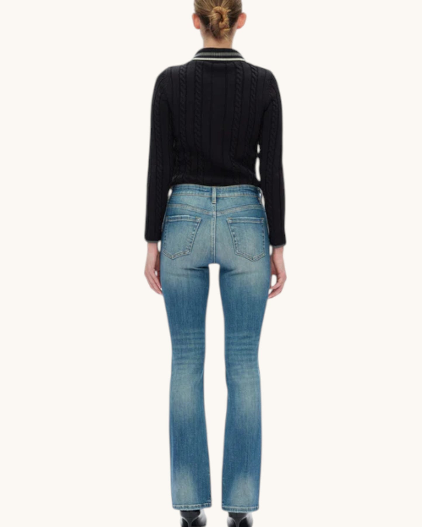 Rear view of a model wearing a black cable-knit sweater with a striped collar and medium-wash blue jeans against a plain off-white background.