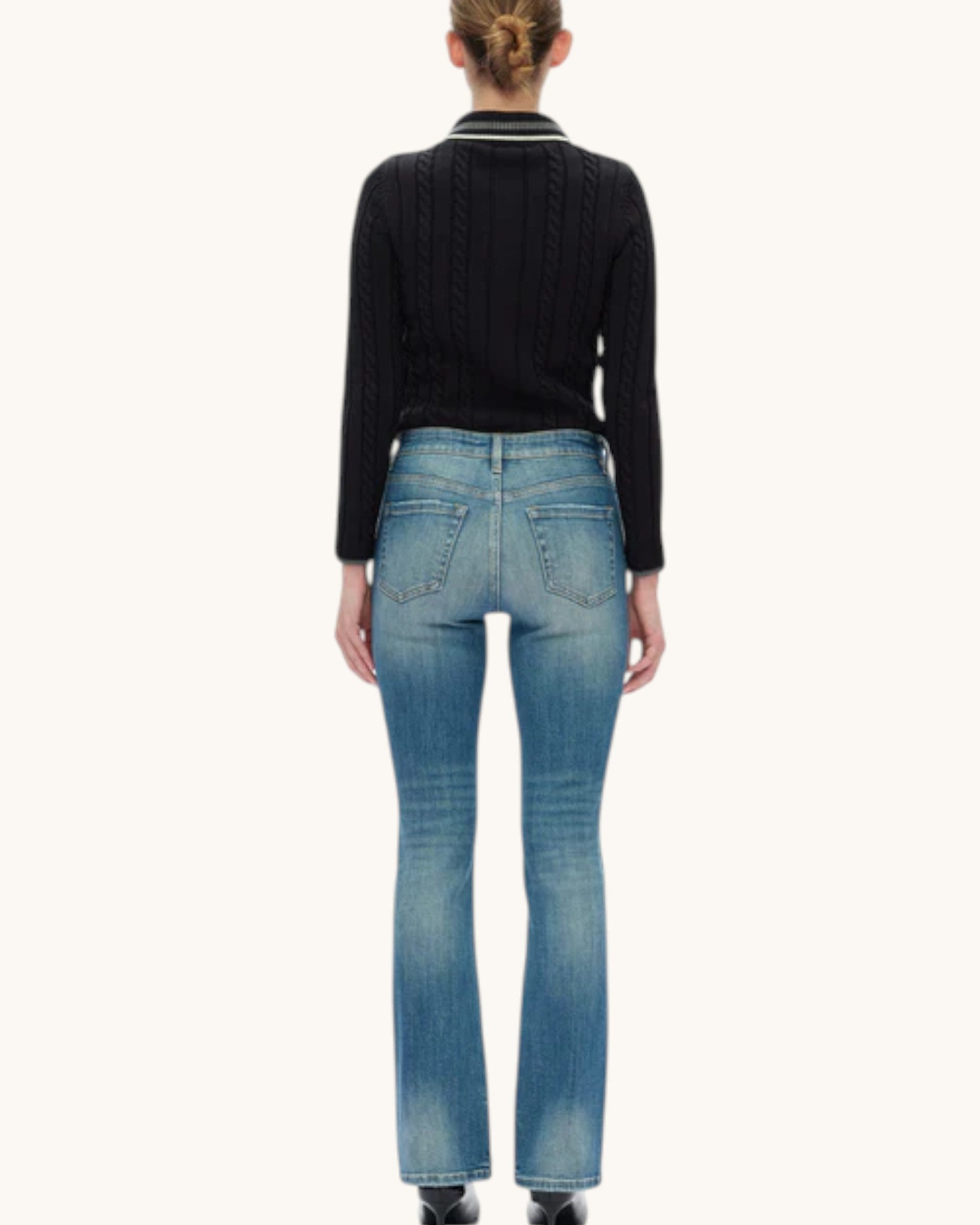 Rear view of a model wearing a black cable-knit sweater with a striped collar and medium-wash blue jeans against a plain off-white background.