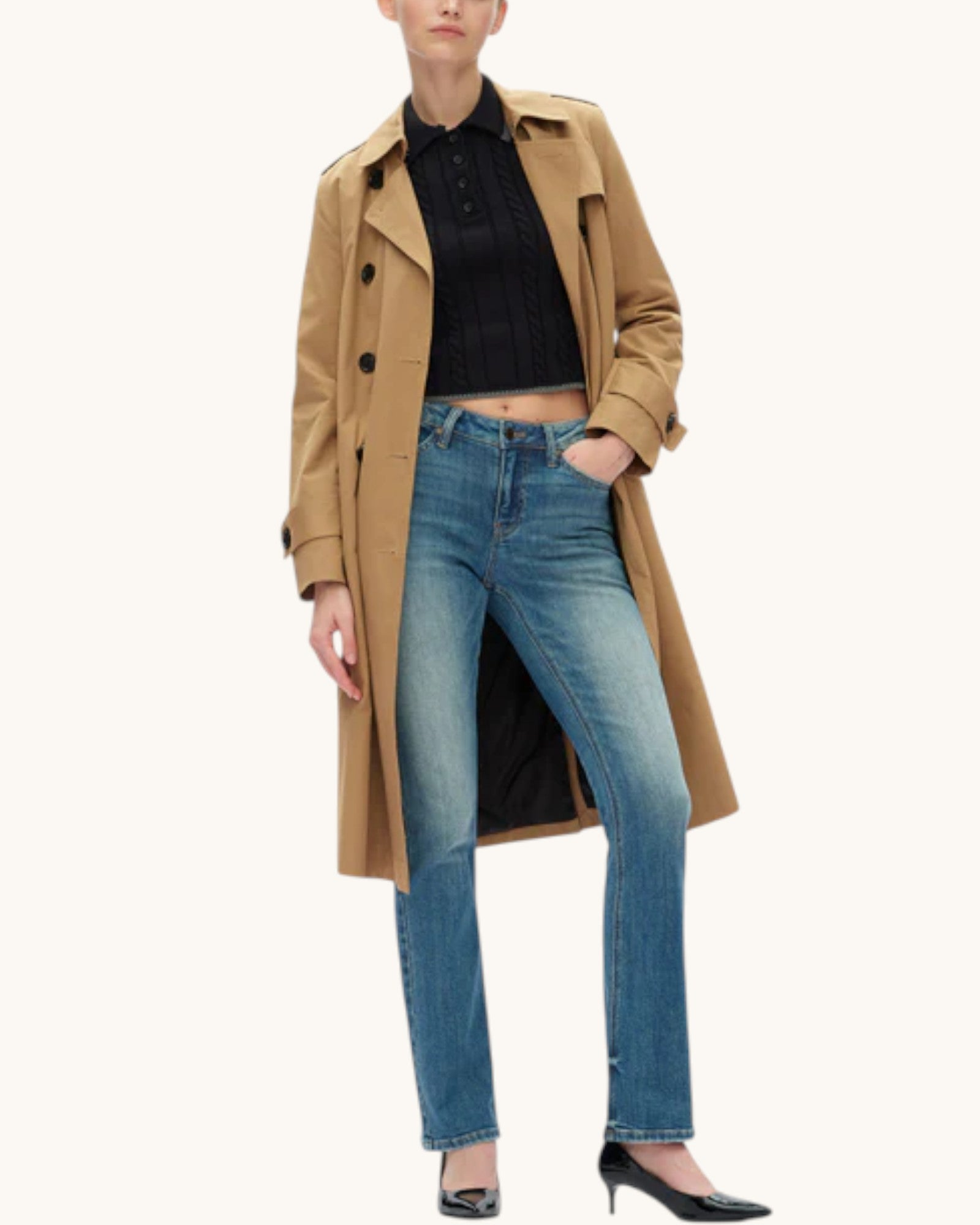 A model wearing a long tan trench coat over a black ribbed top and bootcut blue jeans, paired with black heels, against a plain off-white background.