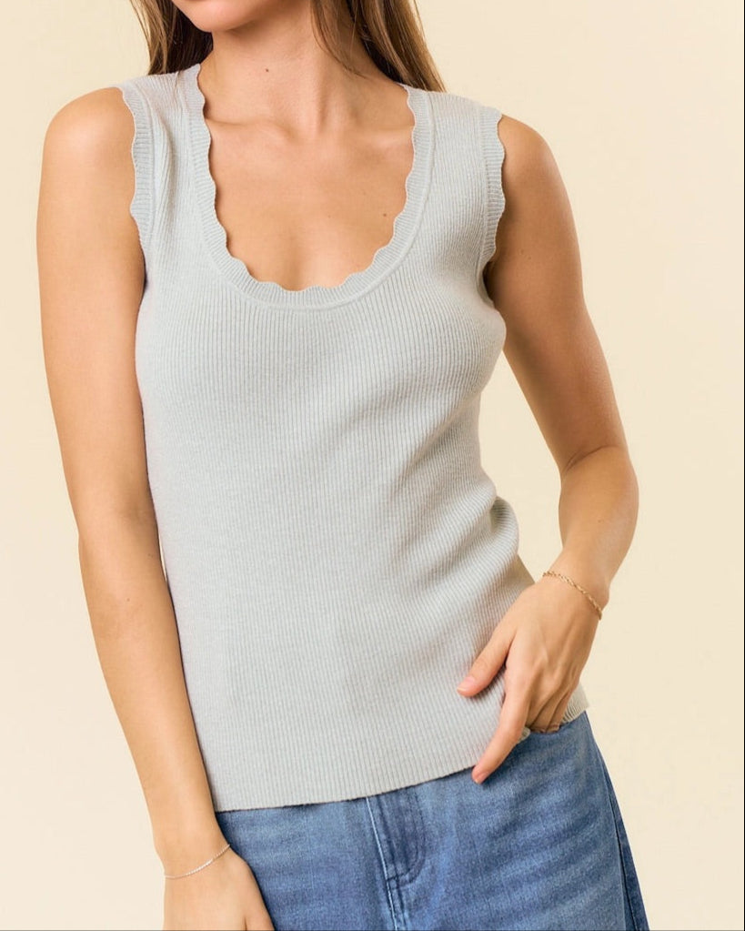 Woman wearing a light green scallop edge and ribbed sleeveless top and blue jeans against a beige background.
