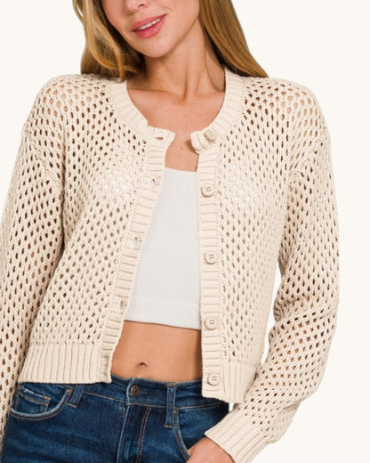 A woman wearing a cream-colored open-knit cropped cardigan over a white crop top and blue jeans.